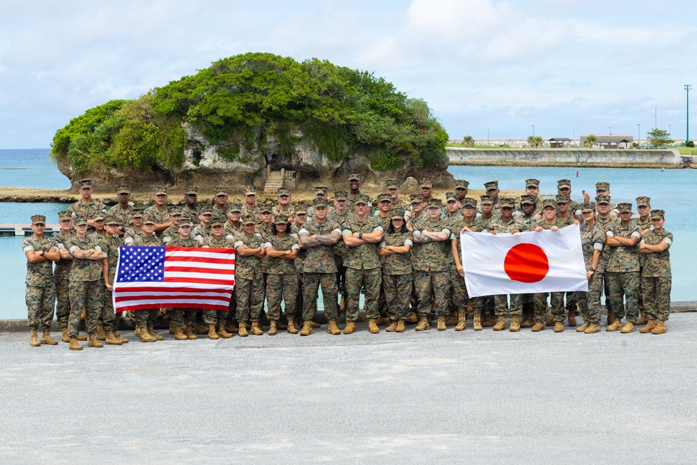 A large group of military personnel in camouflage uniforms pose for a group photograph outdoors near a coastal area. Two flags are prominently displayed: the United States flag on the left and the Japanese flag (Hinomaru) on the right, indicating a joint military exercise or bilateral cooperation event. The backdrop features a distinctive rocky outcrop with vegetation surrounded by turquoise water.