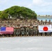 A large group of military personnel in camouflage uniforms pose for a group photograph outdoors near a coastal area. Two flags are prominently displayed: the United States flag on the left and the Japanese flag (Hinomaru) on the right, indicating a joint military exercise or bilateral cooperation event. The backdrop features a distinctive rocky outcrop with vegetation surrounded by turquoise water.