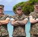 Three U.S. Marines in digital camouflage uniforms (MARPAT) stand with arms crossed in a formal pose against a tropical coastal backdrop. Their name tapes are partially visible, reading 'Quinn' (left), 'Coriano' (center), and a partially obscured name (right). The setting appears to be a tropical island location, likely in the Pacific region.