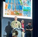 A military personnel in camouflage uniform stands at a clear podium with a microphone, appearing to give a speech or presentation. Two civilians stand behind him, one wearing a red shirt with a dark blazer and another in a dark shirt. Behind them, a large projection screen displays aerial and ground-level construction or development imagery.