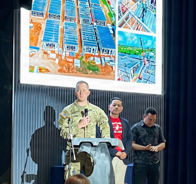 A military personnel in camouflage uniform stands at a clear podium with a microphone, appearing to give a speech or presentation. Two civilians stand behind him, one wearing a red shirt with a dark blazer and another in a dark shirt. Behind them, a large projection screen displays aerial and ground-level construction or development imagery.