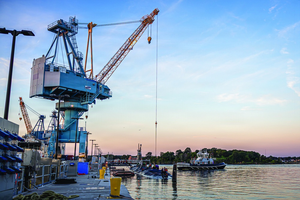 A naval shipyard or industrial port facility photographed at dusk, featuring a large blue harbor crane with an orange lattice boom extending over the waterway. A tugboat and what appears to be a submarine or low-profile vessel are visible in the water alongside the dock. The warm pink and blue twilight sky provides a dramatic backdrop to the industrial scene.