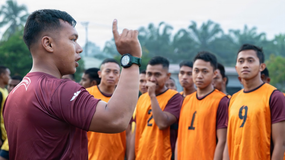A coach or team leader wearing a maroon jersey and a black sports watch is addressing a group of players wearing orange numbered bibs (visible numbers: 1, 2, 9). He is gesturing with his right hand raised, appearing to give tactical instructions or a pre-game briefing. The setting appears to be an outdoor sports field with trees and a light pole visible in the background.