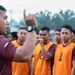 A coach or team leader wearing a maroon jersey and a black sports watch is addressing a group of players wearing orange numbered bibs (visible numbers: 1, 2, 9). He is gesturing with his right hand raised, appearing to give tactical instructions or a pre-game briefing. The setting appears to be an outdoor sports field with trees and a light pole visible in the background.
