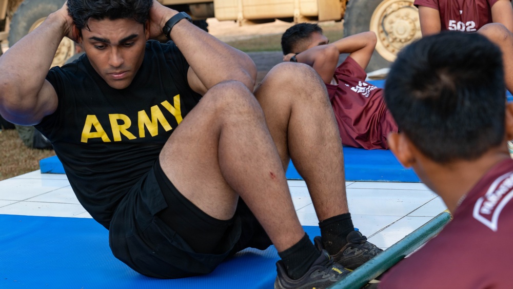 A soldier wearing a black 'ARMY' t-shirt is performing sit-ups on a blue exercise mat outdoors. At least two other individuals in maroon athletic wear are visible in the background performing the same exercise. The setting appears to be a military physical fitness test or training session, with military vehicles visible in the background.