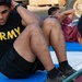 A soldier wearing a black 'ARMY' t-shirt is performing sit-ups on a blue exercise mat outdoors. At least two other individuals in maroon athletic wear are visible in the background performing the same exercise. The setting appears to be a military physical fitness test or training session, with military vehicles visible in the background.