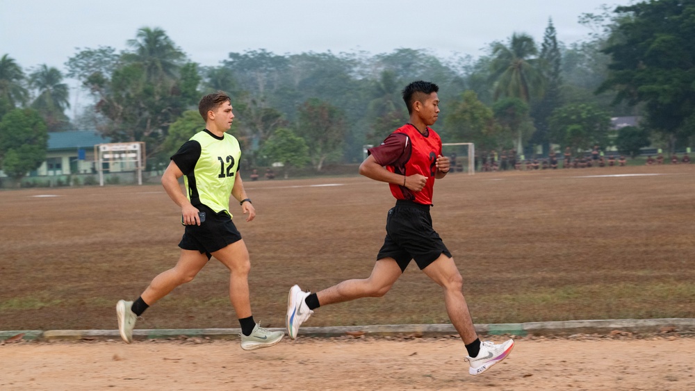 Two male athletes are running on a dirt sports field, likely during a training session or fitness test. One wears a yellow-green bib numbered '12' over a black jersey, while the other wears a red jersey. The background shows a soccer goal, palm trees, and a group of people gathered in the distance.