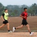 Two male athletes are running on a dirt sports field, likely during a training session or fitness test. One wears a yellow-green bib numbered '12' over a black jersey, while the other wears a red jersey. The background shows a soccer goal, palm trees, and a group of people gathered in the distance.