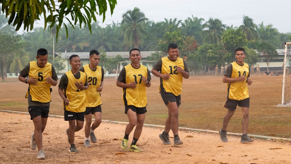 A group of six young men wearing yellow numbered sports bibs over dark military-style clothing are jogging together on a dirt track surrounding a sports field. They appear to be in good spirits, with several smiling. The setting appears to be a military or training facility in a tropical region, evidenced by palm trees and institutional buildings in the background.