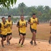 A group of six young men wearing yellow numbered sports bibs over dark military-style clothing are jogging together on a dirt track surrounding a sports field. They appear to be in good spirits, with several smiling. The setting appears to be a military or training facility in a tropical region, evidenced by palm trees and institutional buildings in the background.