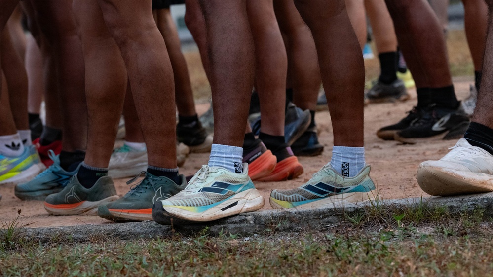 The image shows a close-up view of multiple people's lower legs and feet standing on a dirt surface near a grass edge. The individuals are wearing a variety of athletic running shoes from different brands. The scene appears to be a group gathering, likely before or after a running event or race.