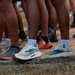 The image shows a close-up view of multiple people's lower legs and feet standing on a dirt surface near a grass edge. The individuals are wearing a variety of athletic running shoes from different brands. The scene appears to be a group gathering, likely before or after a running event or race.