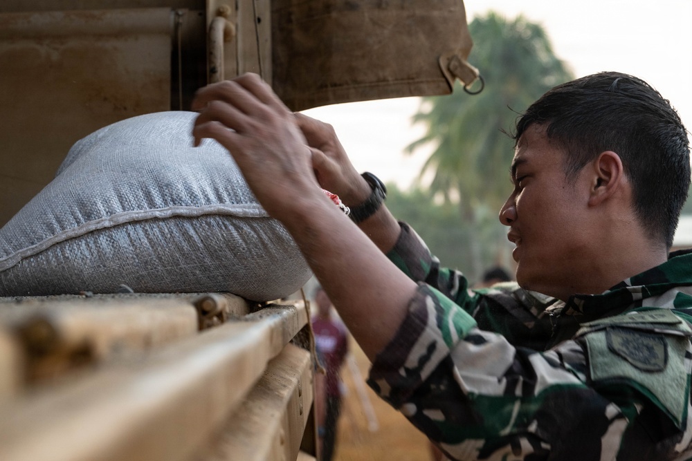 A soldier in camouflage uniform is loading or unloading a large woven sack onto or from a military vehicle. The scene appears to be a relief or logistics operation, likely involving the distribution of supplies such as rice or grain. The setting appears to be an outdoor area with palm trees visible in the background.