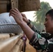 A soldier in camouflage uniform is loading or unloading a large woven sack onto or from a military vehicle. The scene appears to be a relief or logistics operation, likely involving the distribution of supplies such as rice or grain. The setting appears to be an outdoor area with palm trees visible in the background.