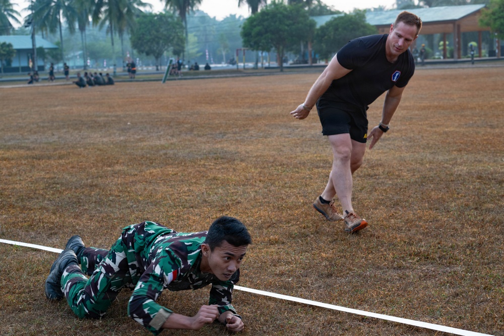 Two individuals are engaged in a military physical training exercise on a grass field. One person in Indonesian camouflage uniform is performing a low crawl under a rope or line, while a second person in a black t-shirt and shorts appears to be supervising or participating alongside. Additional personnel are visible in the background, suggesting a larger group training session.
