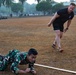 Two individuals are engaged in a military physical training exercise on a grass field. One person in Indonesian camouflage uniform is performing a low crawl under a rope or line, while a second person in a black t-shirt and shorts appears to be supervising or participating alongside. Additional personnel are visible in the background, suggesting a larger group training session.