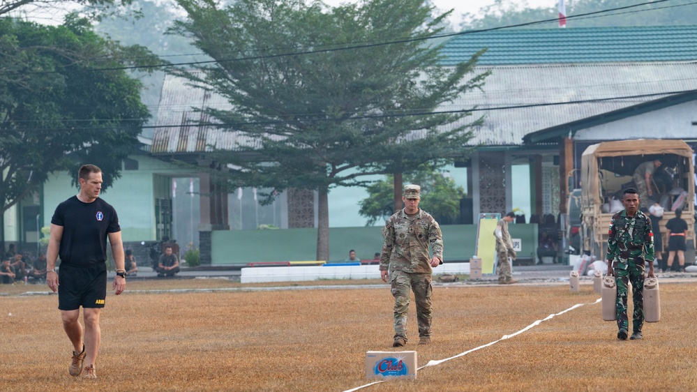 Three military personnel are walking across a dry grass field that appears to be a training or parade ground. The group includes what appears to be a U.S. Army soldier in PT (physical training) attire, a U.S. soldier in full MultiCam/OCP camouflage uniform, and a soldier in green camouflage uniform likely from an Asian military force. The setting suggests a joint military exercise or competition event, with additional personnel and military vehicles visible in the background.