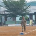 Three military personnel are walking across a dry grass field that appears to be a training or parade ground. The group includes what appears to be a U.S. Army soldier in PT (physical training) attire, a U.S. soldier in full MultiCam/OCP camouflage uniform, and a soldier in green camouflage uniform likely from an Asian military force. The setting suggests a joint military exercise or competition event, with additional personnel and military vehicles visible in the background.