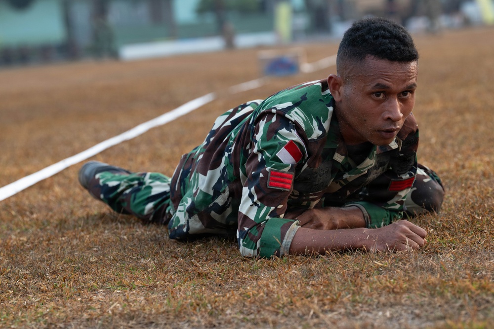 A soldier in Indonesian military camouflage uniform is performing a low crawl exercise on a dry grass field. He is lying prone and moving forward under what appears to be a rope or wire obstacle course boundary. The soldier displays visible rank insignia and a red-and-white Indonesian flag patch on his uniform.