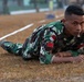 A soldier in Indonesian military camouflage uniform is performing a low crawl exercise on a dry grass field. He is lying prone and moving forward under what appears to be a rope or wire obstacle course boundary. The soldier displays visible rank insignia and a red-and-white Indonesian flag patch on his uniform.