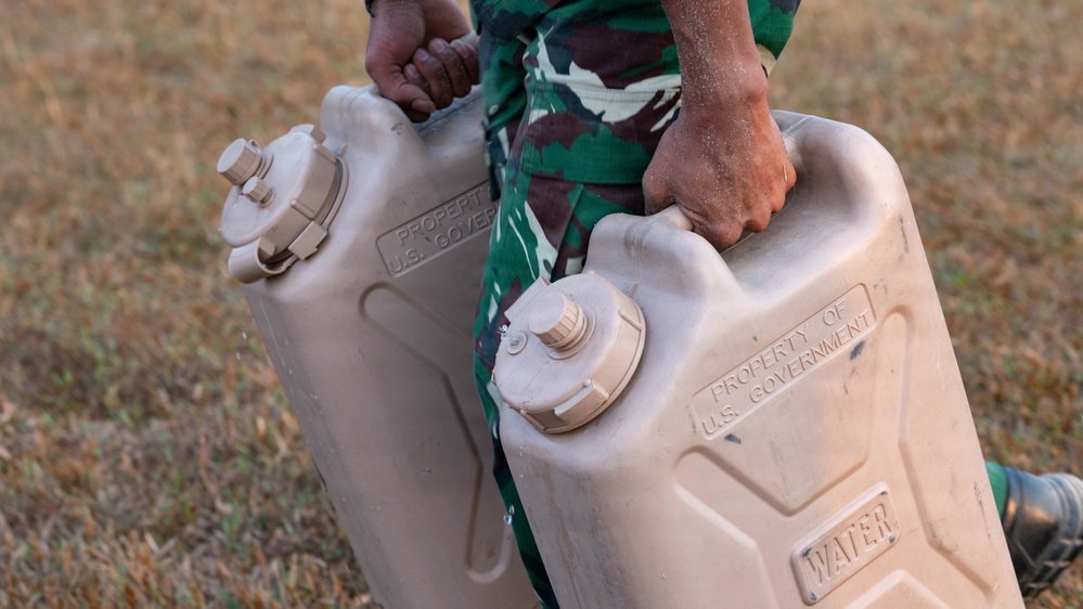 A person wearing green camouflage military uniform is carrying two large plastic water jerry cans. Both containers are embossed with 'PROPERTY OF U.S. GOVERNMENT' and labeled 'WATER', indicating military-issue equipment. The scene appears to be outdoors on dry, grassy terrain.