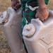 A person wearing green camouflage military uniform is carrying two large plastic water jerry cans. Both containers are embossed with 'PROPERTY OF U.S. GOVERNMENT' and labeled 'WATER', indicating military-issue equipment. The scene appears to be outdoors on dry, grassy terrain.