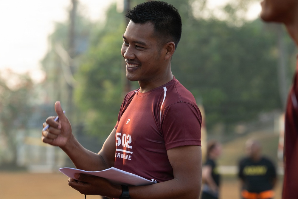 A smiling young man wearing a dark red/maroon athletic shirt with '502' and partial text visible is holding papers and a pen outdoors. He appears to be in a coaching or officiating role at what looks like a sports field or court. The image is taken during golden hour lighting with a blurred background showing trees and other people.