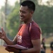 A smiling young man wearing a dark red/maroon athletic shirt with '502' and partial text visible is holding papers and a pen outdoors. He appears to be in a coaching or officiating role at what looks like a sports field or court. The image is taken during golden hour lighting with a blurred background showing trees and other people.