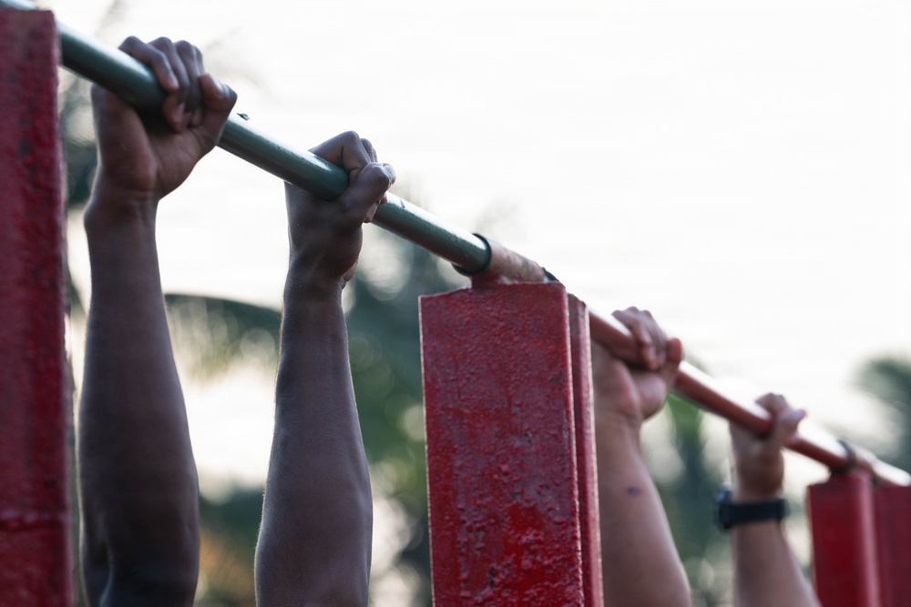 The image shows multiple pairs of hands gripping a horizontal pull-up bar mounted on red metal support structures outdoors. Several individuals appear to be performing or preparing for pull-up exercises simultaneously. The photo is taken from a low angle with a shallow depth of field, creating a bokeh background of trees and sky.