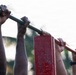 The image shows multiple pairs of hands gripping a horizontal pull-up bar mounted on red metal support structures outdoors. Several individuals appear to be performing or preparing for pull-up exercises simultaneously. The photo is taken from a low angle with a shallow depth of field, creating a bokeh background of trees and sky.