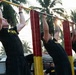 Three individuals wearing matching black 'ARMY' t-shirts are performing pull-ups on a red and yellow painted outdoor pull-up bar structure. The setting appears to be a military physical training area surrounded by palm trees, suggesting a tropical location. A fourth person in an Army shirt is visible in the background observing.