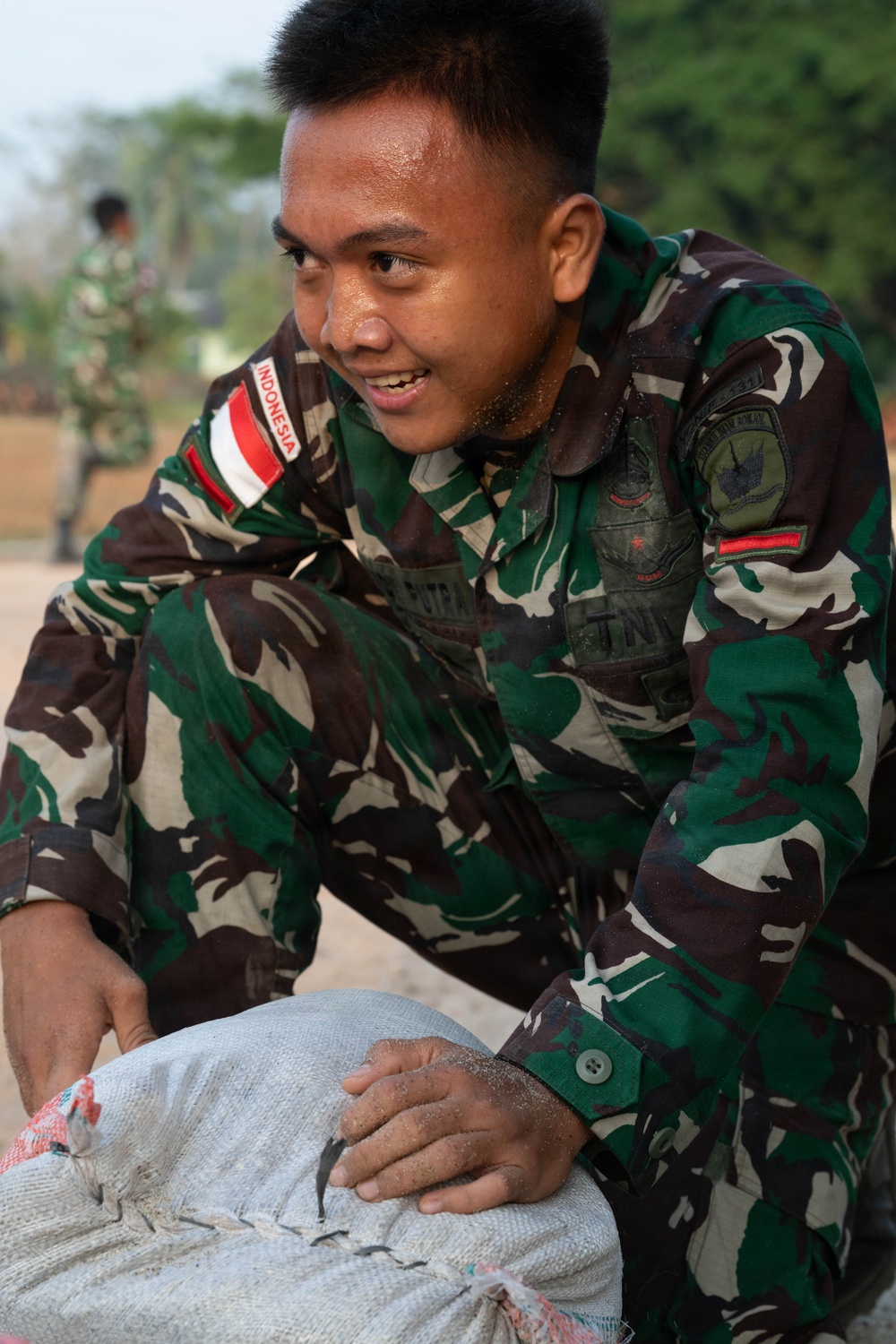 A soldier wearing Indonesian military (TNI - Tentara Nasional Indonesia) camouflage uniform is handling a large sack or sandbag, appearing to be engaged in a physical task or exercise. The soldier is smiling and visibly sweating, suggesting strenuous physical activity. A second soldier in similar uniform is visible in the blurred background.