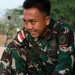 A soldier wearing Indonesian military (TNI - Tentara Nasional Indonesia) camouflage uniform is handling a large sack or sandbag, appearing to be engaged in a physical task or exercise. The soldier is smiling and visibly sweating, suggesting strenuous physical activity. A second soldier in similar uniform is visible in the blurred background.