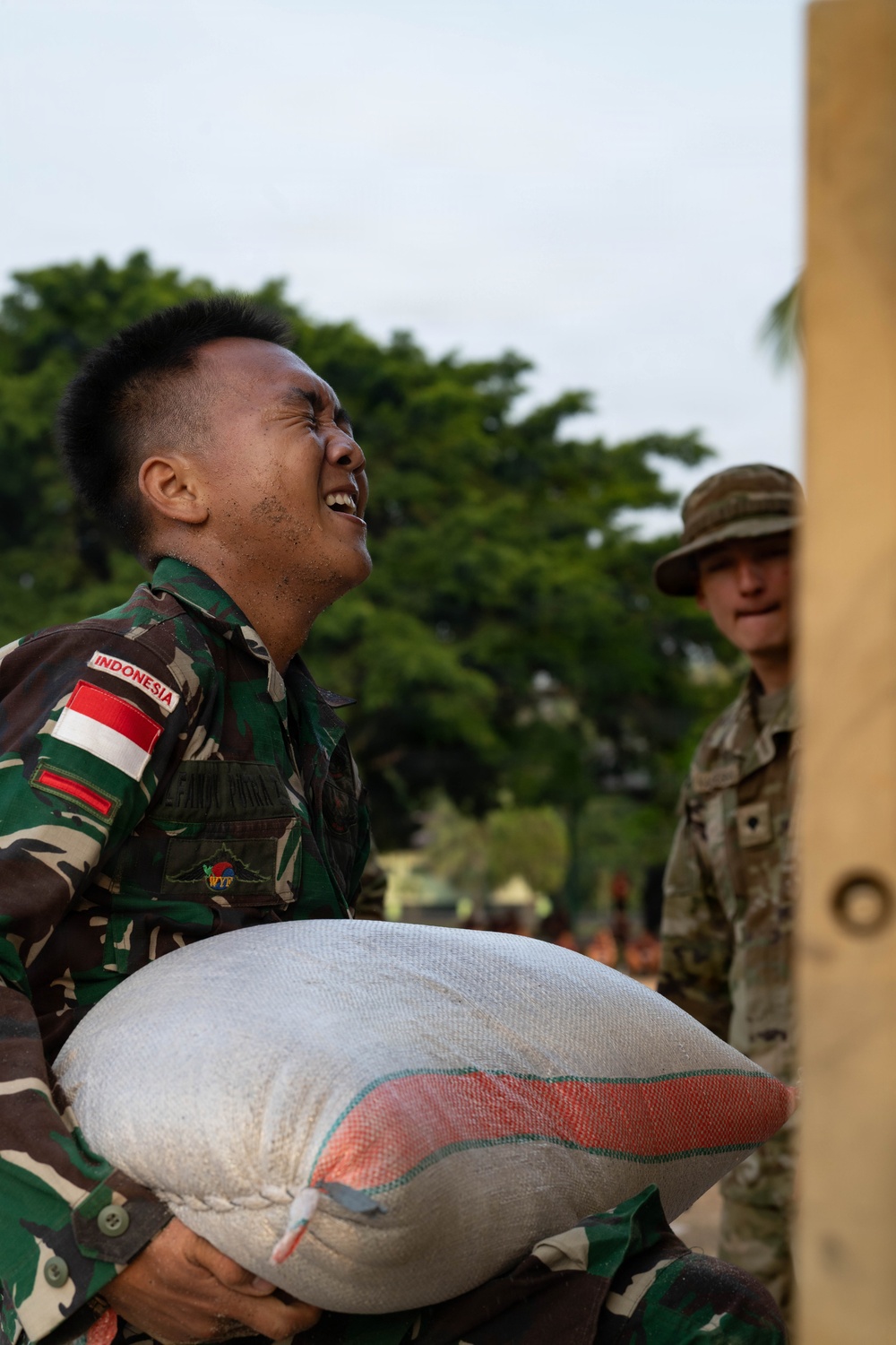 An Indonesian military soldier is straining under the weight of a large heavy sack, his face showing visible exertion and effort. A second soldier in a different camouflage uniform and bush hat stands in the background observing. The setting appears to be an outdoor area with trees visible, suggesting a joint military exercise or humanitarian aid operation.