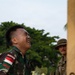 An Indonesian military soldier is straining under the weight of a large heavy sack, his face showing visible exertion and effort. A second soldier in a different camouflage uniform and bush hat stands in the background observing. The setting appears to be an outdoor area with trees visible, suggesting a joint military exercise or humanitarian aid operation.