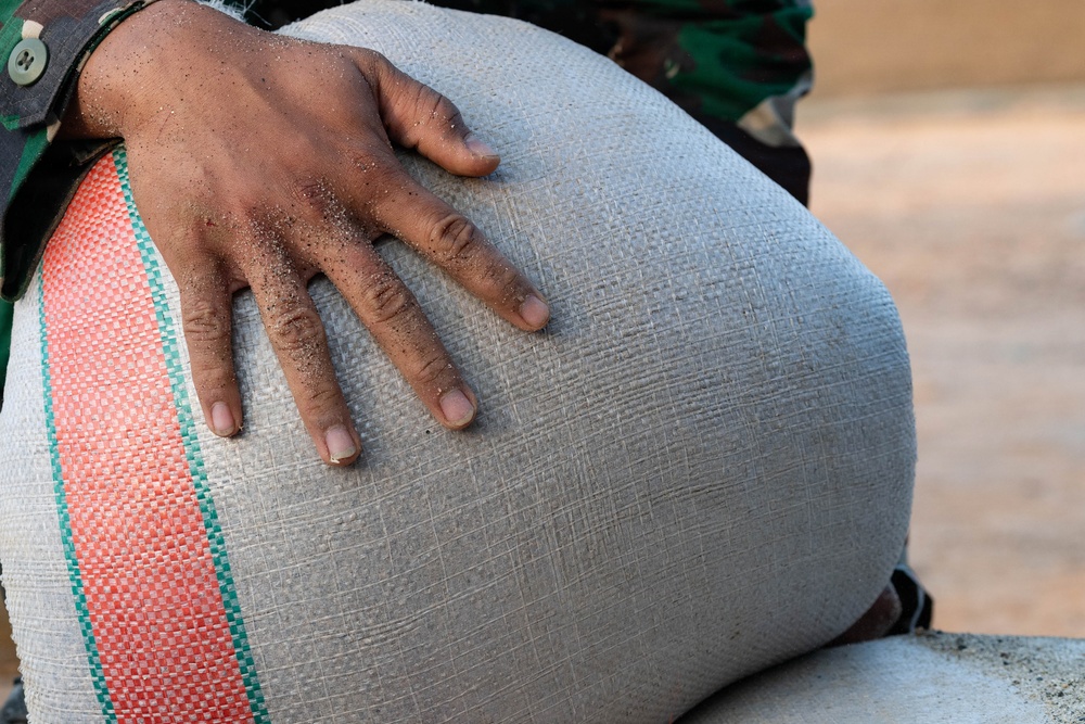 A close-up photograph showing a dark-skinned hand resting on a large woven sack or bag. The hand appears dusty or covered with fine particles, suggesting manual labor. The person is wearing a camouflage-patterned shirt visible in the upper portion of the frame.