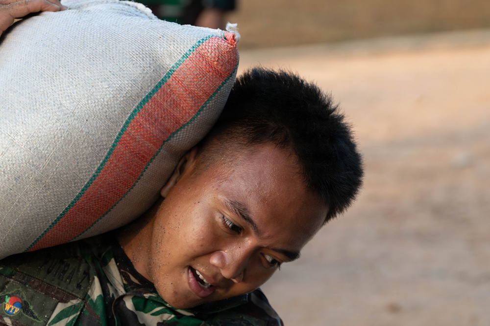 A young man wearing camouflage military or work uniform is carrying a large heavy sack on his shoulder and neck. He appears to be straining under the weight, with his mouth open and body bent forward. The sack is woven fabric with red and green striped trim, consistent with grain or rice sacks.