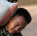 A young man wearing camouflage military or work uniform is carrying a large heavy sack on his shoulder and neck. He appears to be straining under the weight, with his mouth open and body bent forward. The sack is woven fabric with red and green striped trim, consistent with grain or rice sacks.