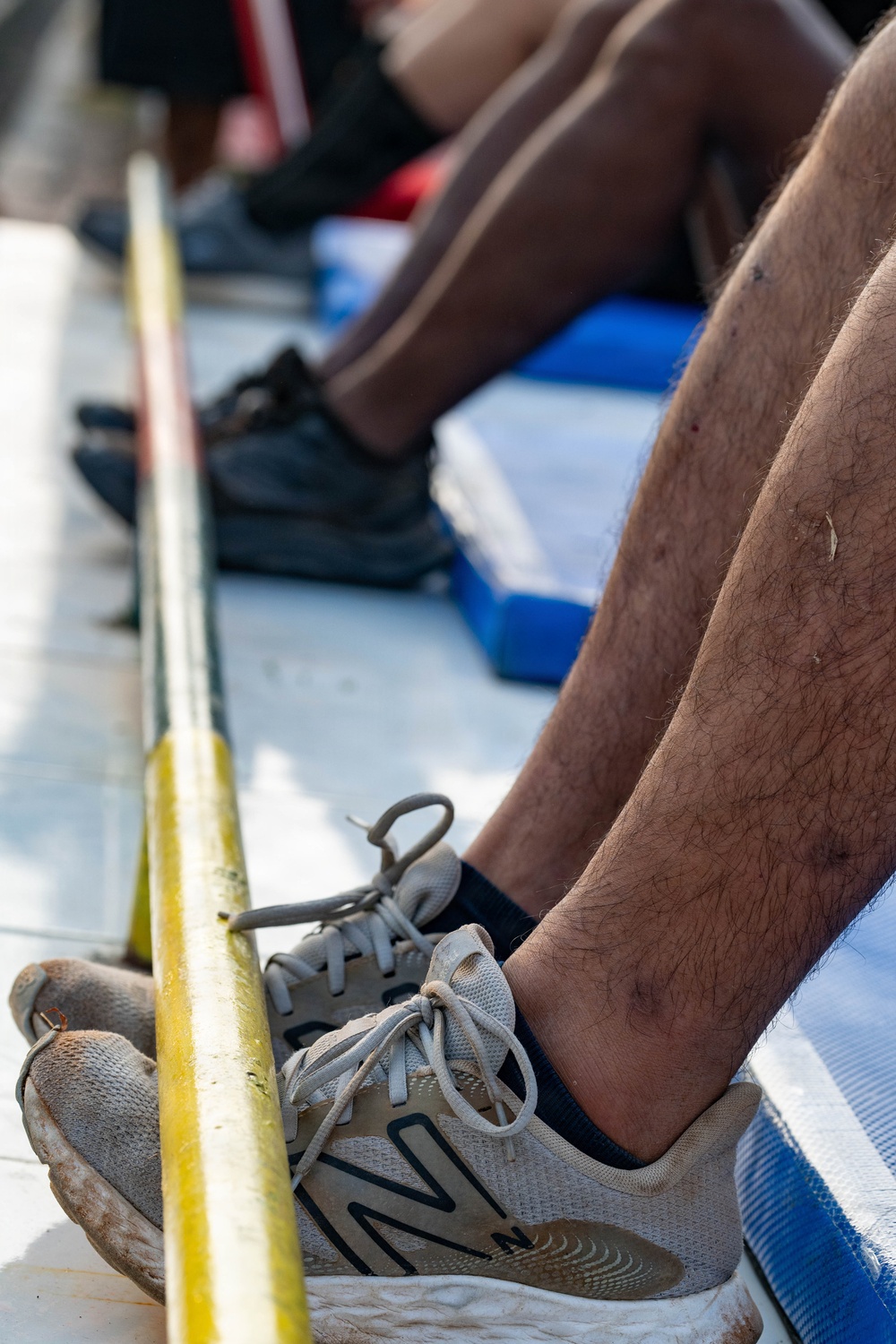 A close-up photograph showing the lower legs and feet of multiple individuals seated in what appears to be a rowing boat or similar watercraft. The foreground features a person wearing worn New Balance sneakers resting against a yellow painted rail or gunwale. Blue seat cushions or foot stretchers are visible beneath the rowers.