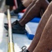 A close-up photograph showing the lower legs and feet of multiple individuals seated in what appears to be a rowing boat or similar watercraft. The foreground features a person wearing worn New Balance sneakers resting against a yellow painted rail or gunwale. Blue seat cushions or foot stretchers are visible beneath the rowers.