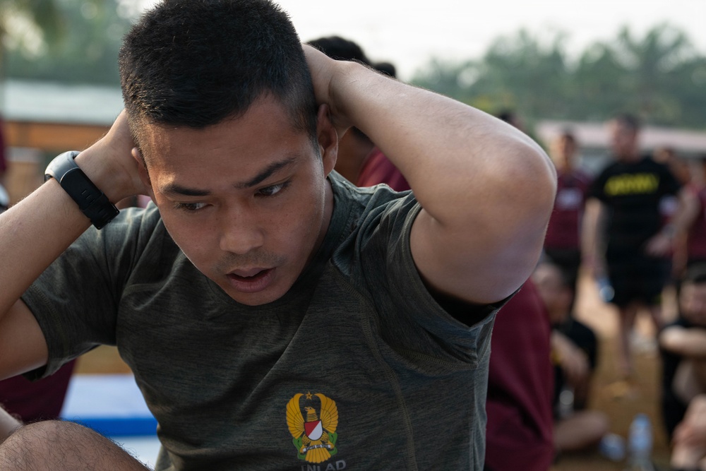 A young male soldier is performing sit-ups or crunches with his hands clasped behind his head during what appears to be a military physical fitness test or training exercise. He is wearing an olive green t-shirt bearing a military insignia, likely Indonesian Armed Forces (TNI/SALAD emblem). The background shows other participants in various colored shirts engaged in similar activities in an outdoor setting.