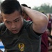 A young male soldier is performing sit-ups or crunches with his hands clasped behind his head during what appears to be a military physical fitness test or training exercise. He is wearing an olive green t-shirt bearing a military insignia, likely Indonesian Armed Forces (TNI/SALAD emblem). The background shows other participants in various colored shirts engaged in similar activities in an outdoor setting.