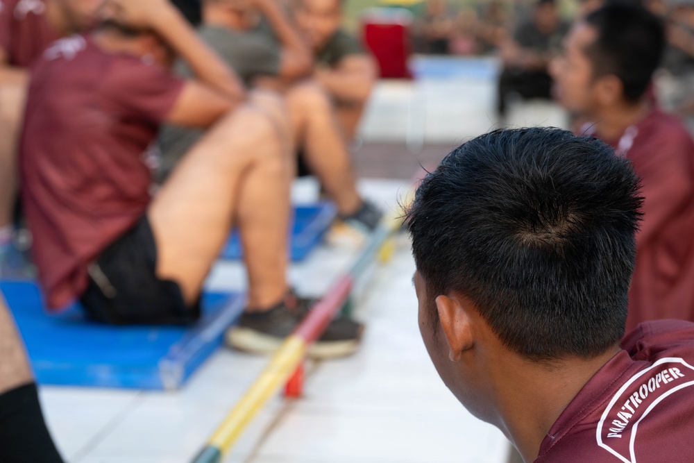 The image shows a group of individuals wearing matching maroon/burgundy shirts, appearing to be engaged in a physical fitness or training activity. The foreground features the back of a person's head with text on their shirt reading 'PARATROOPER'. Several people in the background are performing what appears to be sit-ups or crunches on blue exercise mats.
