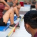 The image shows a group of individuals wearing matching maroon/burgundy shirts, appearing to be engaged in a physical fitness or training activity. The foreground features the back of a person's head with text on their shirt reading 'PARATROOPER'. Several people in the background are performing what appears to be sit-ups or crunches on blue exercise mats.