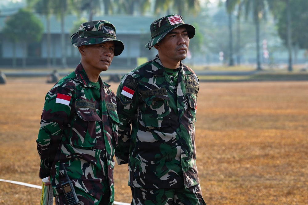 Two Indonesian military personnel (TNI - Tentara Nasional Indonesia) are standing on a dry grass field, appearing to observe something off-camera. Both wear standard Indonesian Army camouflage uniforms with Indonesian flag patches. The soldier on the right has a 'MEDIC' tab on his hat and a name tape reading 'JEPRI', while the left soldier carries a radio.