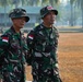 Two Indonesian military personnel (TNI - Tentara Nasional Indonesia) are standing on a dry grass field, appearing to observe something off-camera. Both wear standard Indonesian Army camouflage uniforms with Indonesian flag patches. The soldier on the right has a 'MEDIC' tab on his hat and a name tape reading 'JEPRI', while the left soldier carries a radio.