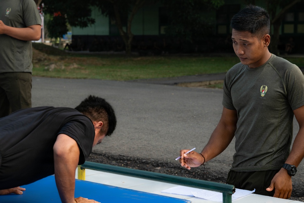 The image shows what appears to be a military physical fitness test being conducted outdoors. One individual in a black shirt is performing push-ups near a low bar or measuring apparatus, while another person in an olive military t-shirt holds a pen and observes or records results. A third person in olive attire is visible in the background.