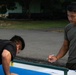 The image shows what appears to be a military physical fitness test being conducted outdoors. One individual in a black shirt is performing push-ups near a low bar or measuring apparatus, while another person in an olive military t-shirt holds a pen and observes or records results. A third person in olive attire is visible in the background.