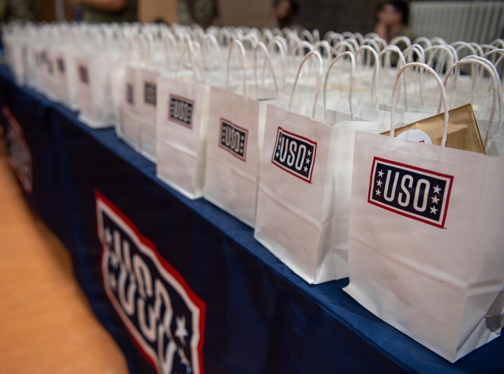 A long table covered with a blue USO-branded tablecloth is lined with numerous white gift bags, each bearing the USO logo in navy blue and red. The bags appear to be prepared for distribution, likely at a military support or appreciation event. The depth of field creates a perspective effect showing the bags extending far into the background.