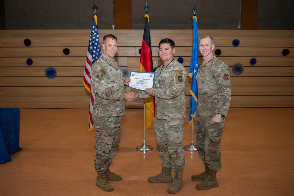 Three U.S. Air Force personnel in OCP (Operational Camouflage Pattern) uniforms are posed during a formal recognition ceremony. The center individual is receiving a certificate, with the name 'Mariano M. Doviardo' and the rank 'Staff Sergeant' visible on the document. The ceremony appears to be taking place at a U.S. military installation in Germany, evidenced by the American, German, and U.S. Air Force flags displayed in the background.